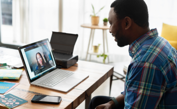 A person works at a wooden table on their laptop and speaks to someone over video conferencing 